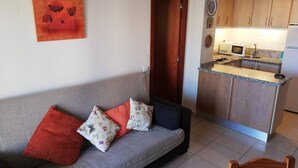 A living room with a grey sofa, red and floral cushions, and a wooden door leading to a kitchen with wooden cabinets and a white refrigerator. - Infante E8-7º Solo Familias! (Playa de Gandía)