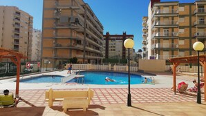 A swimming pool area with a bench, a lifeguard tower, and a multi-story building in the background. - Infante E8-7º Solo Familias! (Playa de Gandía)