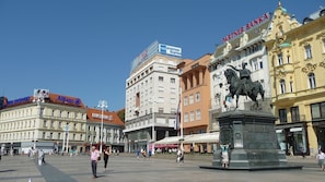 A public square with a statue, surrounded by buildings with various signs, including a bank and a restaurant. - Oval Main Square Apartment (Zagreb)
