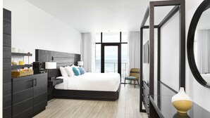 A modern hotel room with a large bed, a dresser, a chair, and a view of the sea. - Wave Resort (Long Branch)