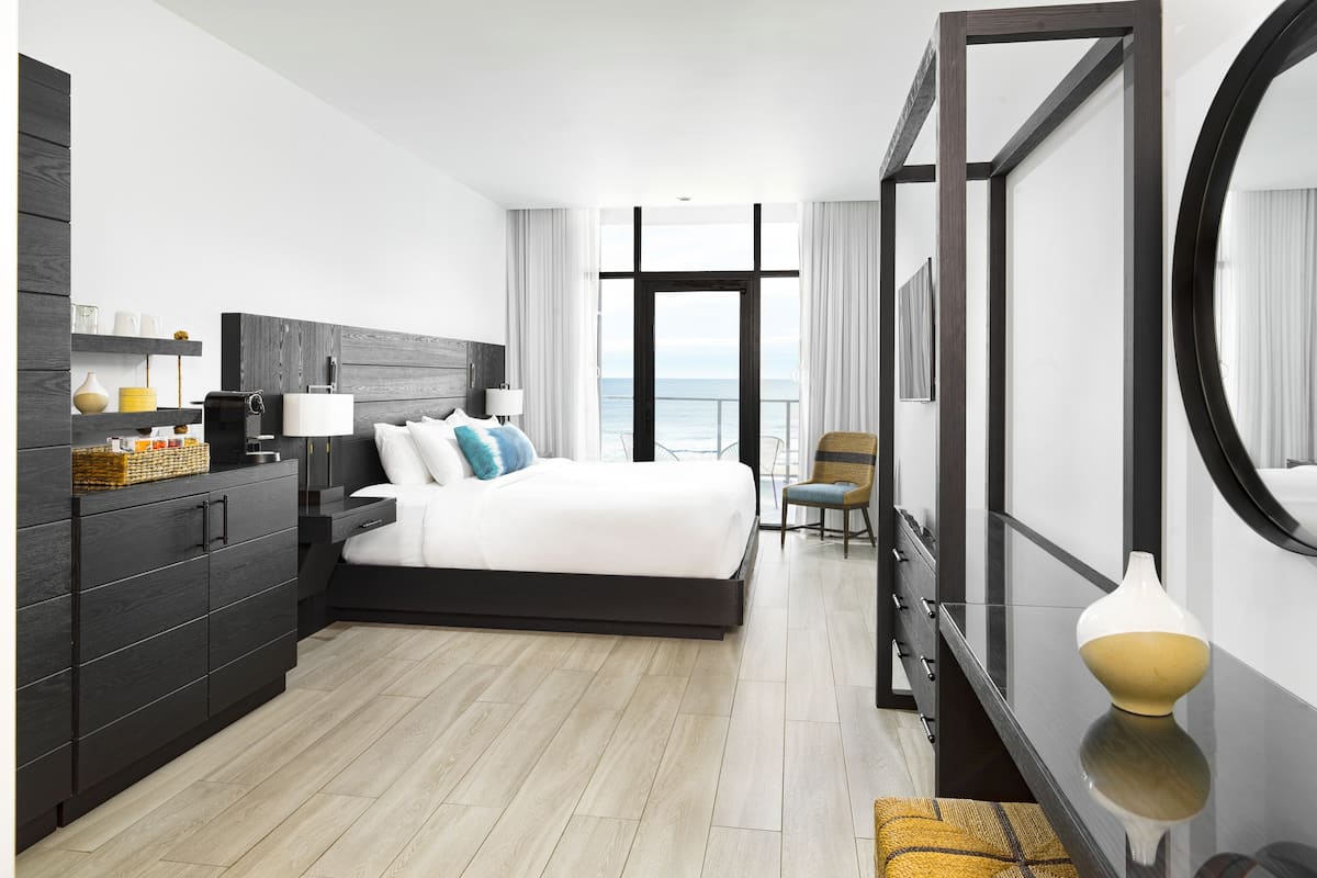 A modern hotel room with a large bed, a dresser, a chair, and a view of the sea.