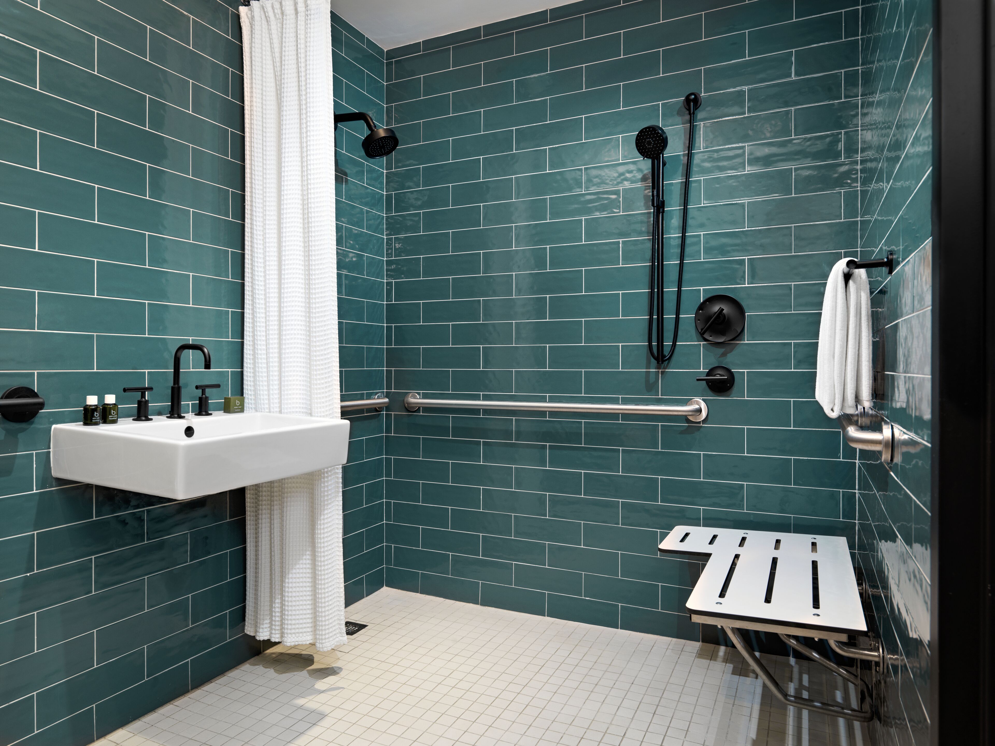 A modern bathroom with teal tiles, a white sink, a handheld showerhead, a grab bar, and a towel rack.