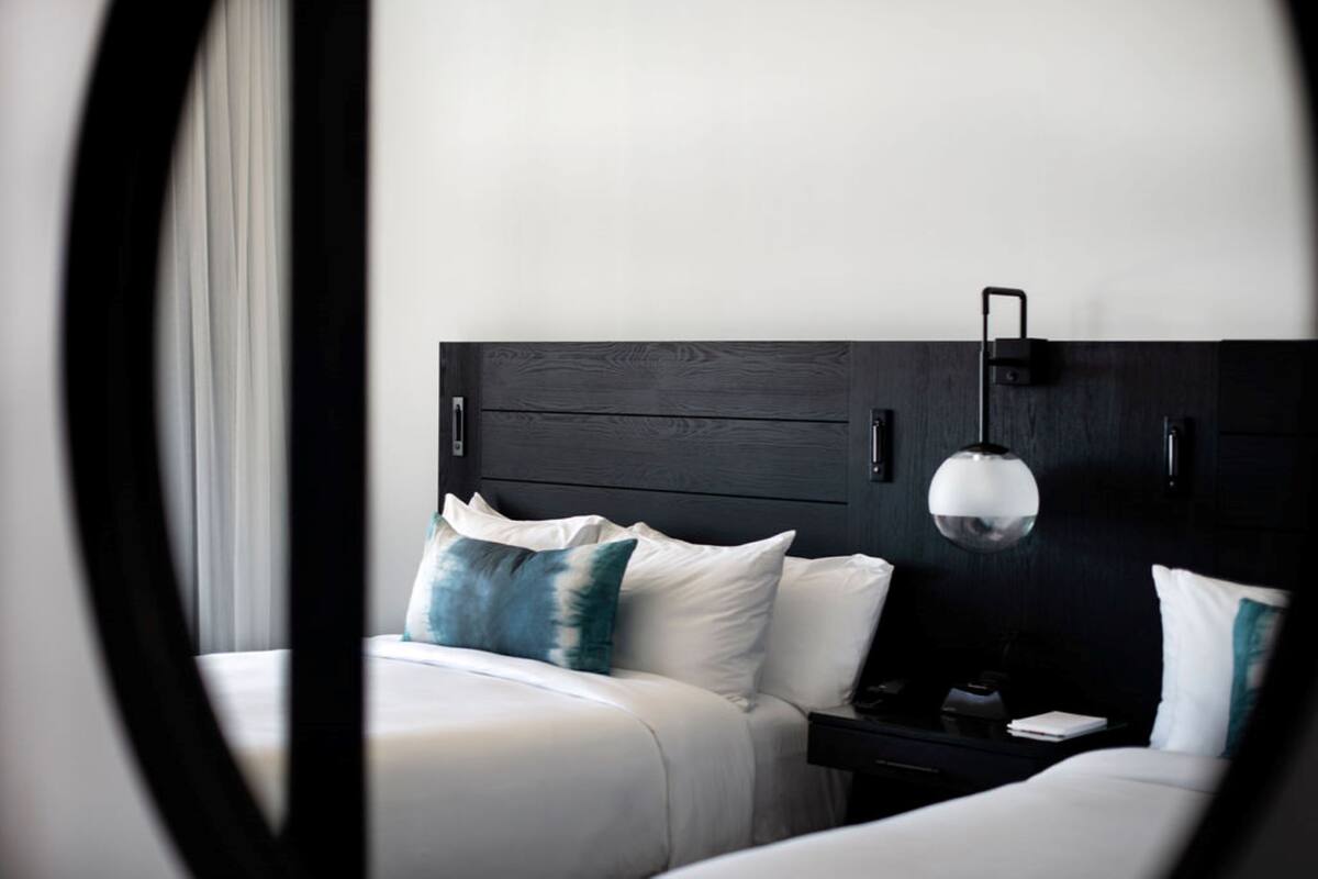 A modern hotel room with a dark wood headboard, white bedding, and a single bedside table with a lamp.