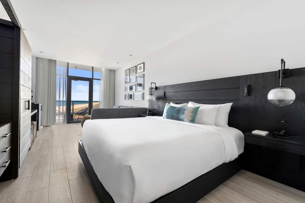 A modern hotel room with a large bed, a desk, and a view of the ocean.