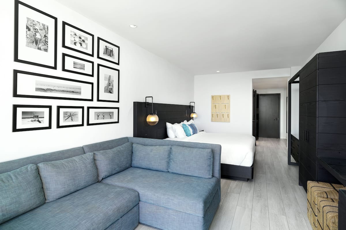 A modern hotel room with a grey sofa, a bed with white bedding, a black wardrobe, and framed pictures on the wall.