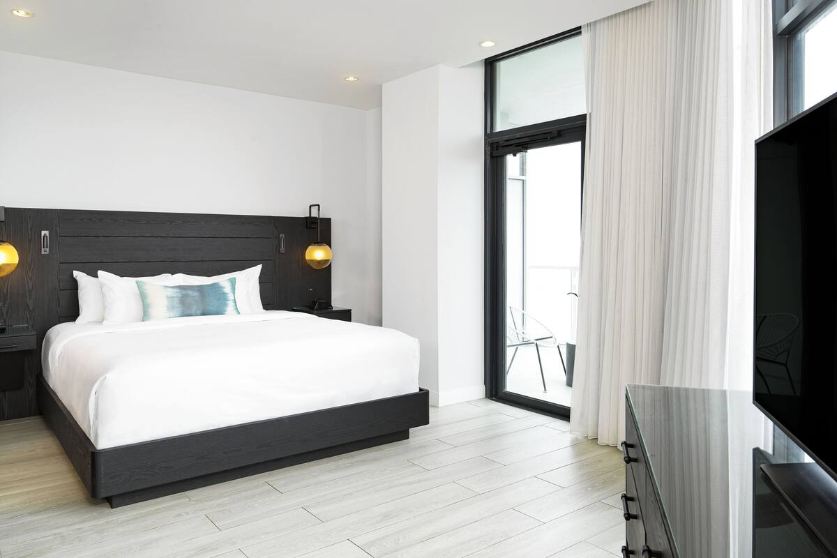 A modern hotel room with a large bed, a TV, a balcony with a chair, and a large window with curtains.