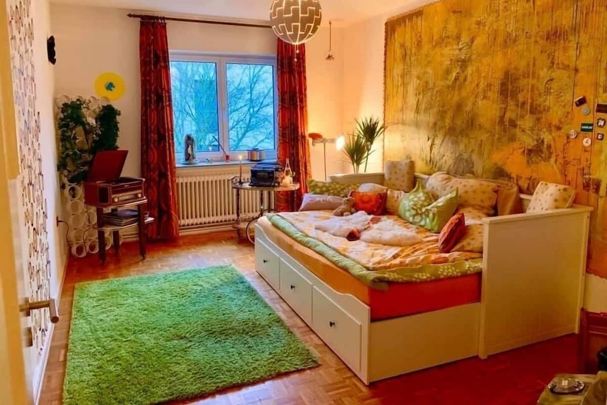 Een slaapkamer met een bed, een bureau, een radiator, een raam en een groene vloerkleed.
