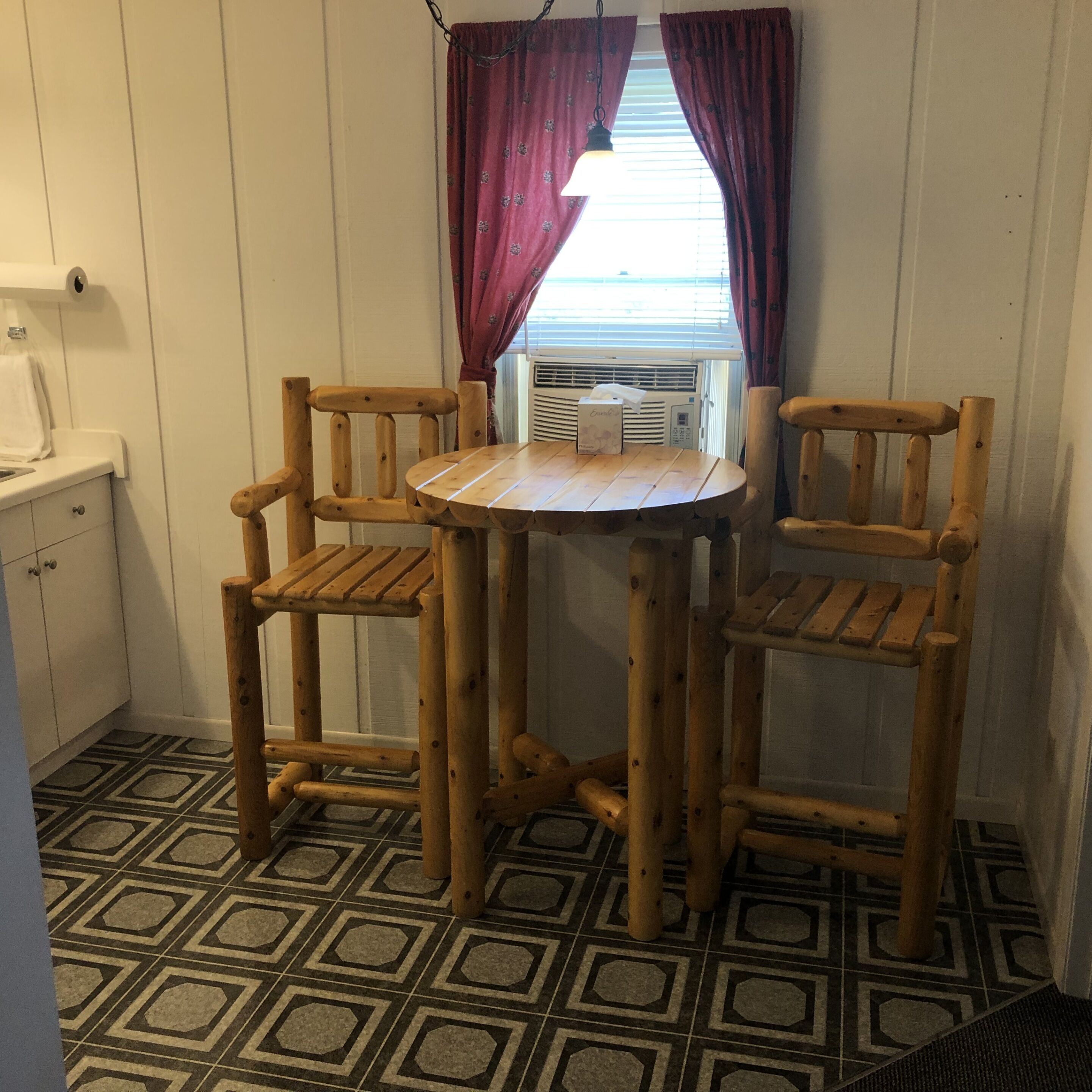 A small kitchen with a round wooden table and two chairs, a window with curtains, and a patterned floor.