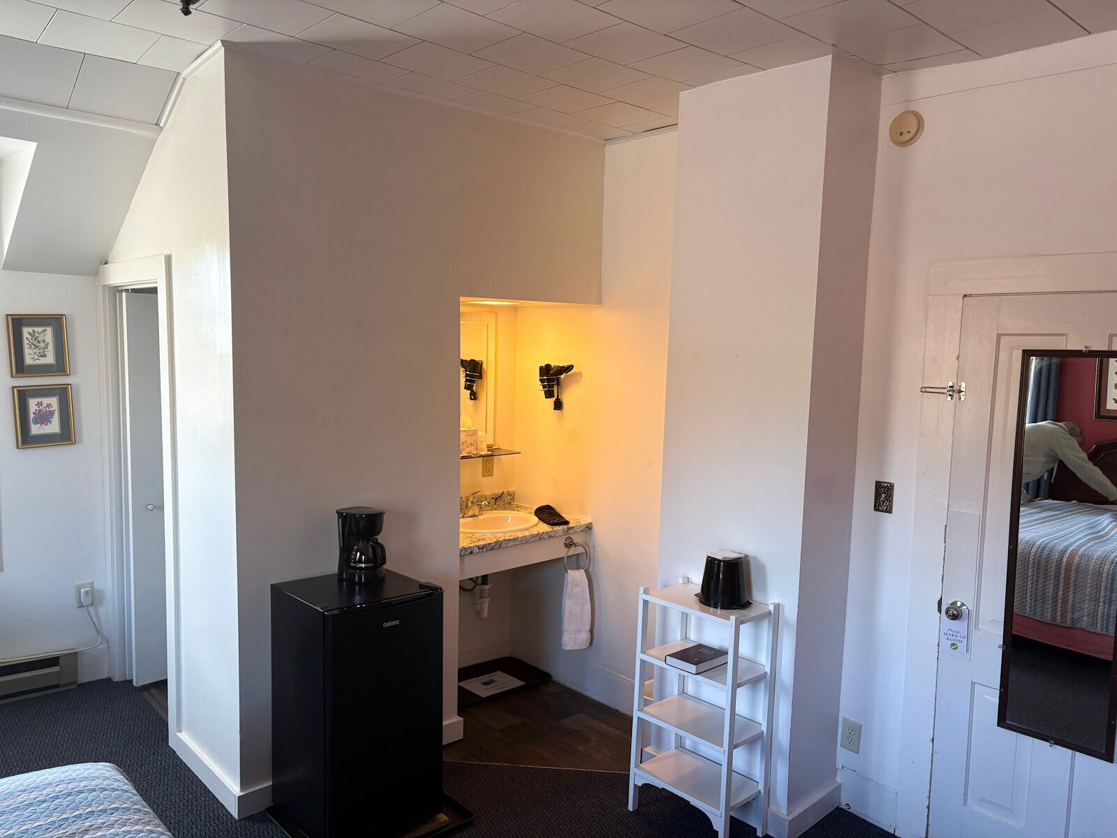 Une chambre d’hôtel compacte comprenant un petit coin cuisine, un lit simple, une porte blanche avec un miroir et une étagère blanche.