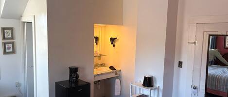 Une chambre d’hôtel compacte comprenant un petit coin cuisine, un lit simple, une porte blanche avec un miroir et une étagère blanche.
