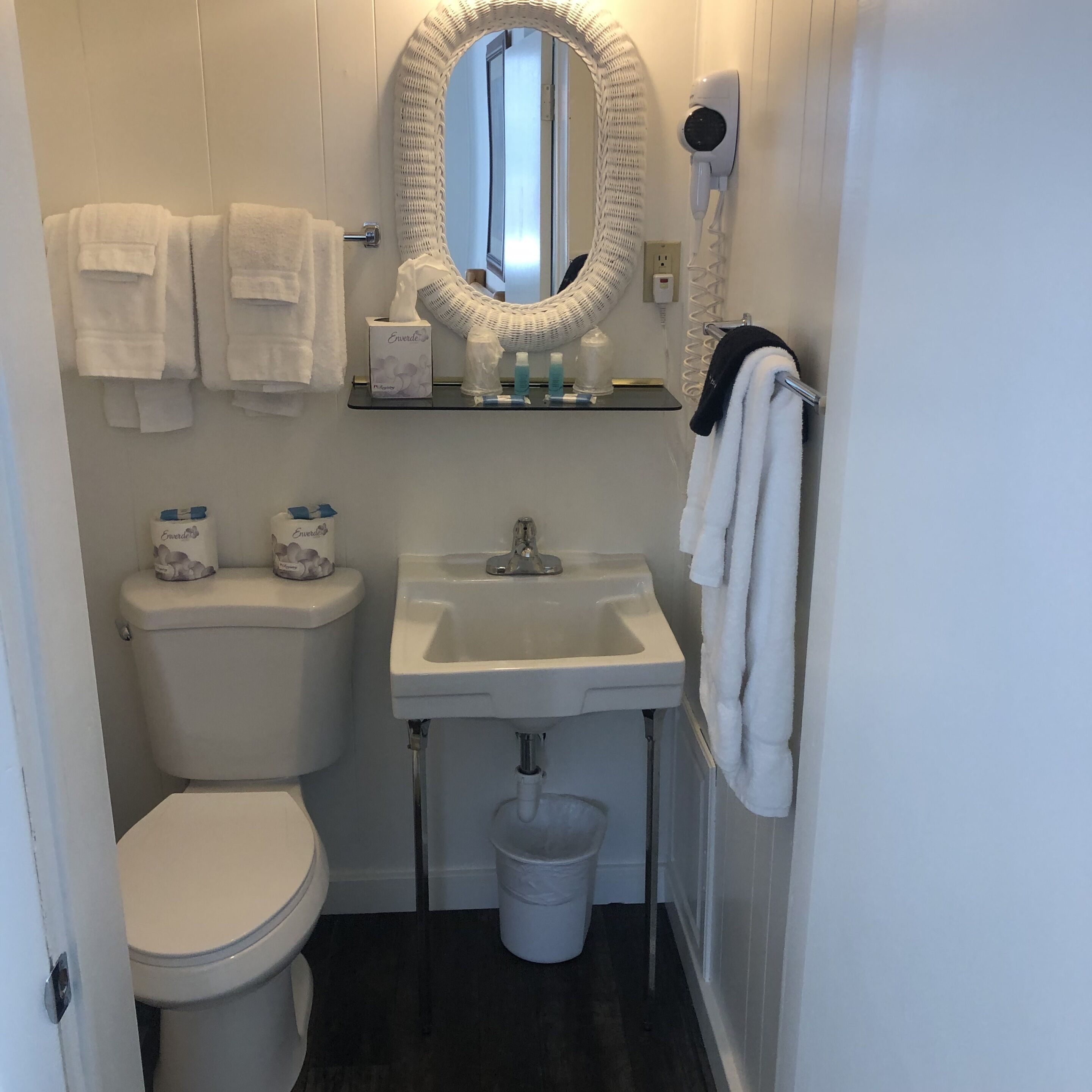 Une petite salle de bain propre, équipée de toilettes, d’un lavabo et d’un miroir. Des serviettes sont accrochées au mur et un sèche-cheveux est fixé au mur.
