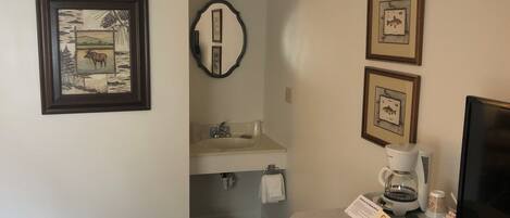 A hotel room with a bathroom, a sink, a mirror, framed pictures on the wall, a coffee maker, and a TV.