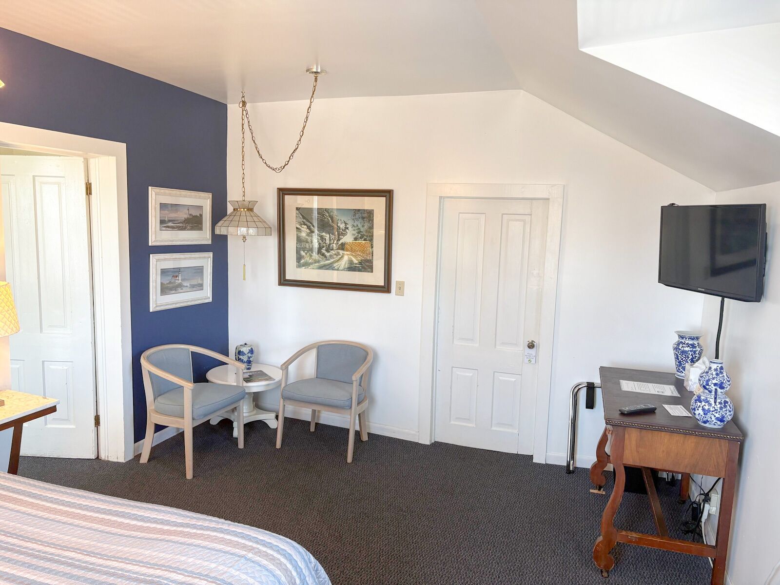 A room with a bed, two chairs, a small table, a TV, and artwork on the wall.