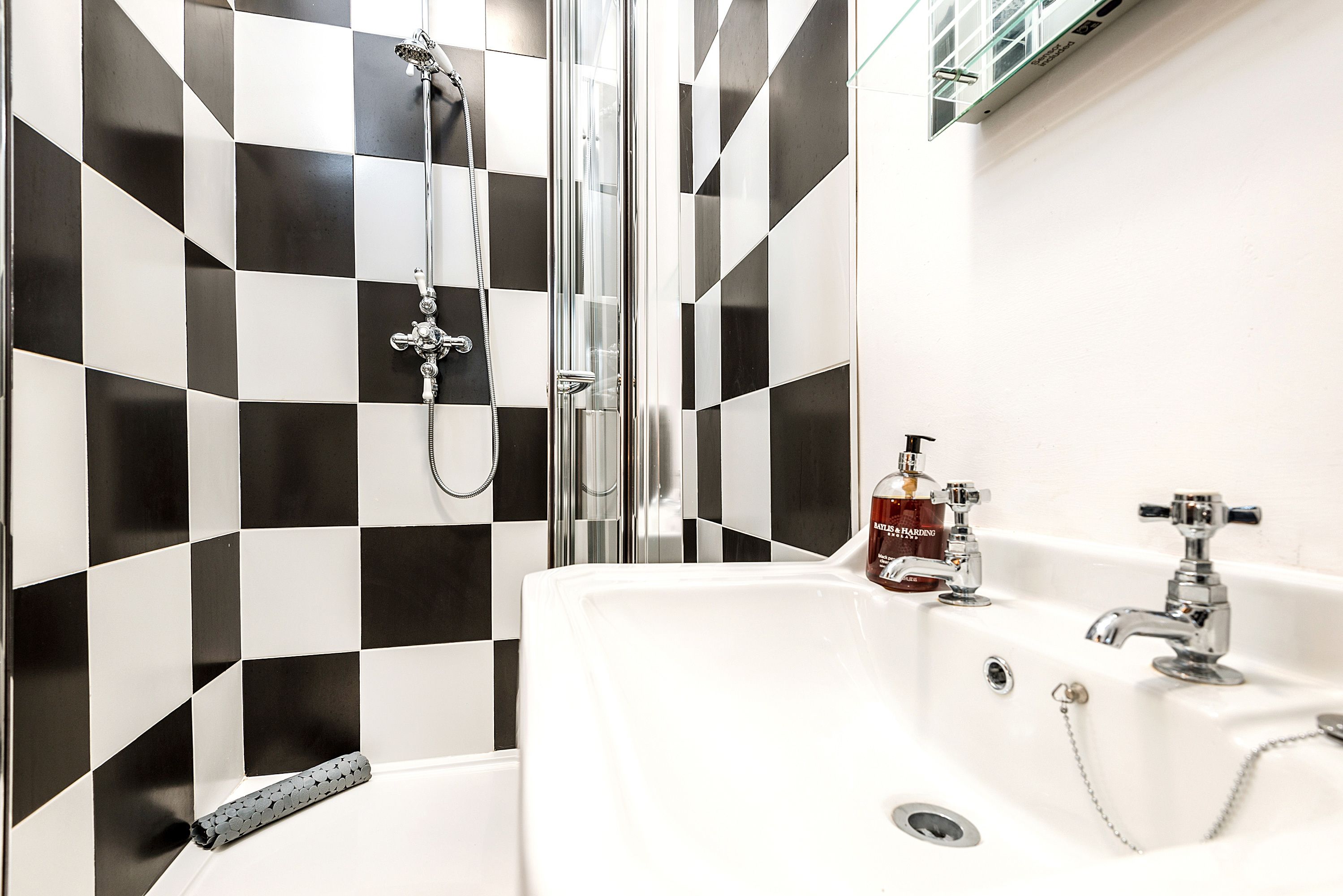 A bathroom with a checkered black and white tile wall, a glass-enclosed shower, a white sink with a chrome faucet, and a bottle of hand soap on the counter.