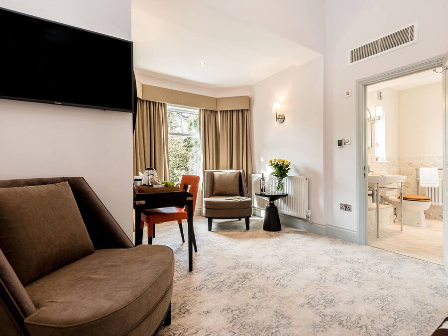 Ein Hotelzimmer mit einem an der Wand montierten Flachbildfernseher, einem Sofa, einem Esstisch mit Stühlen, einem kleinen Beistelltisch mit einer Vase und einem durch eine offene Tür einsehbaren Badezimmer.