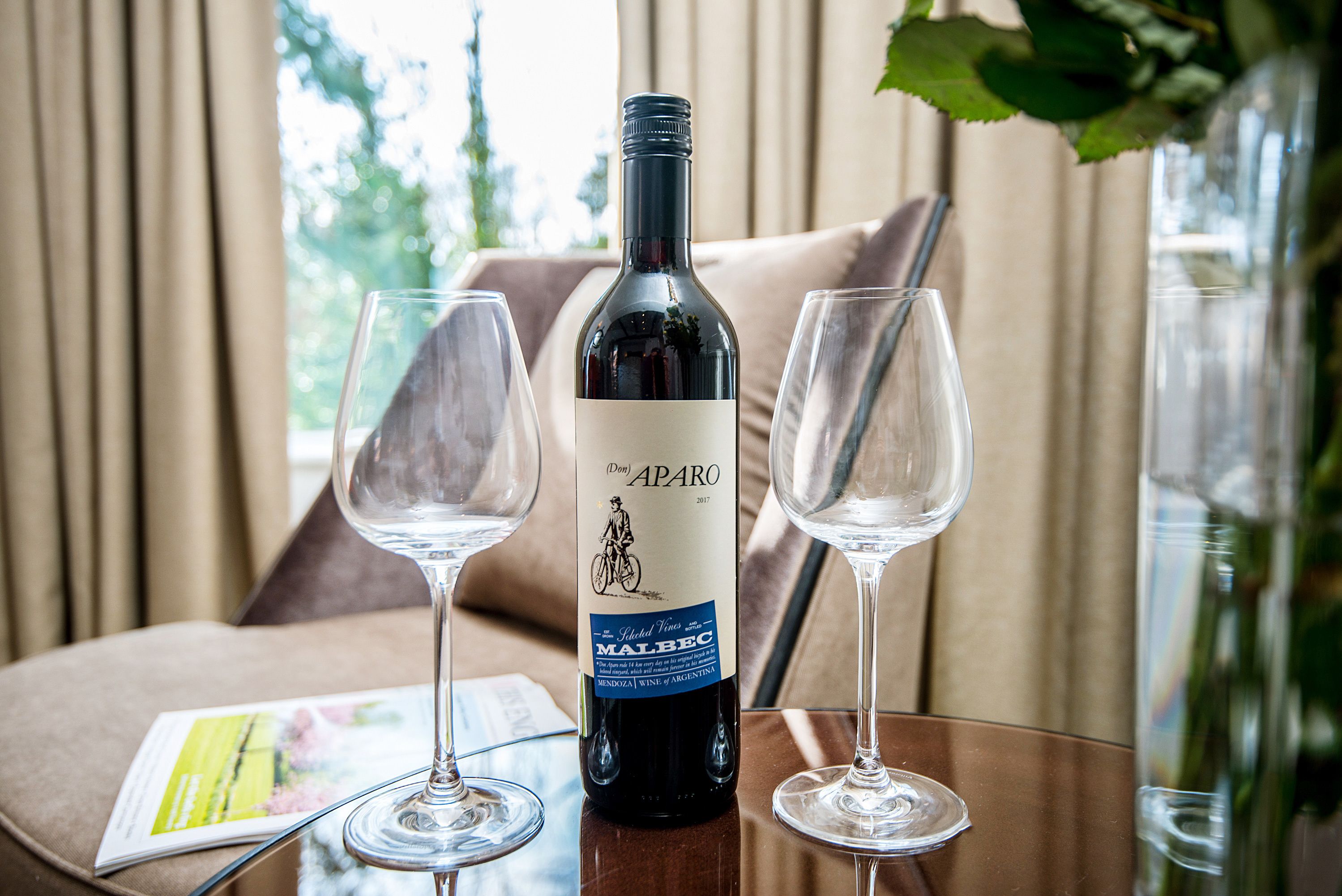 A wine bottle labeled "APARO" and two empty wine glasses on a table with a magazine and a vase with flowers.