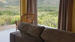 Una habitación con un sofá y una silla de mimbre, un ventanal con cortinas y vistas a un paisaje montañoso.