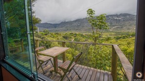 Una terraza de madera con una mesa y sillas, con vista a un paisaje montañoso, vegetación y cielo nublado.