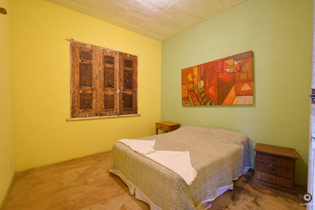 A bedroom with a bed, a wooden window, a small wooden nightstand, and a painting on the wall.