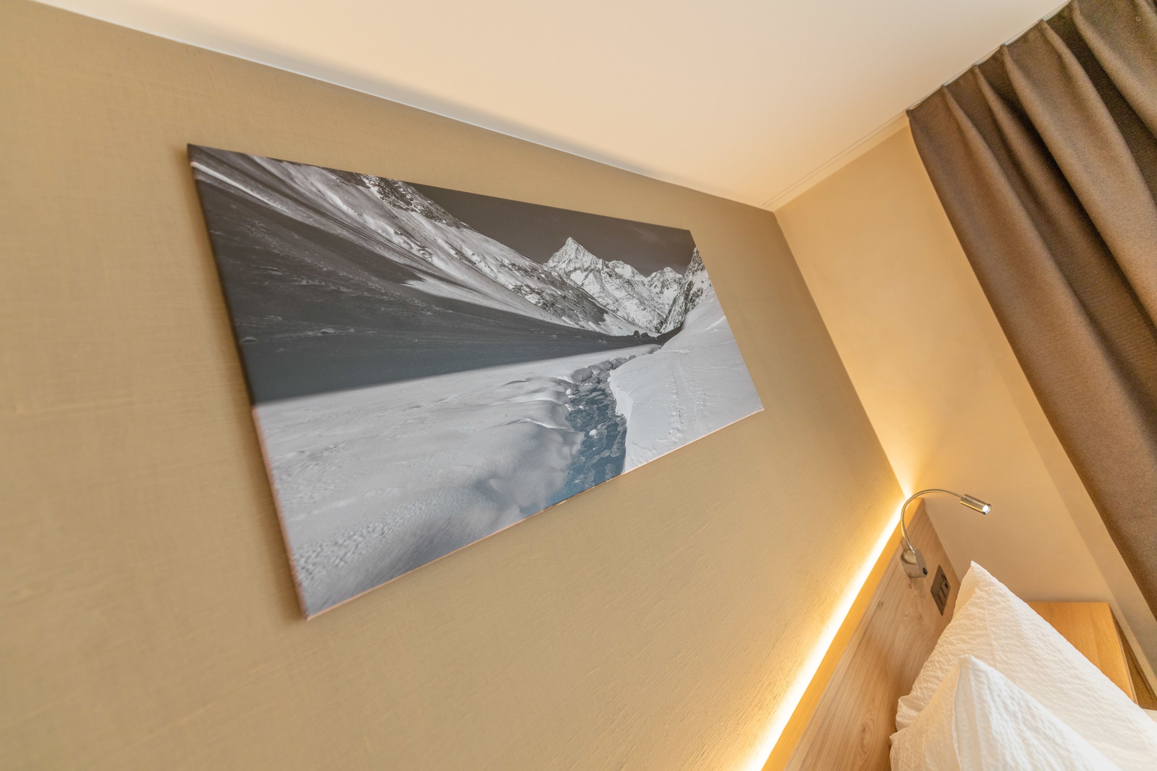 Ein Hotelzimmer mit großem Landschaftsbild von Bergen an der Wand, einem Bett mit weißer Bettwäsche und einem Holz-Kopfteil.