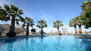 A clear blue swimming pool surrounded by palm trees, with a view of a body of water and buildings in the background.