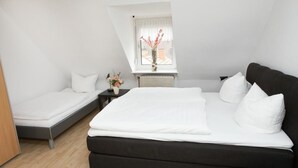 A bedroom with a large bed, a smaller bed, a nightstand with a vase of flowers, and a window with a view of the outside. - Zwehrener Hof (Kassel)