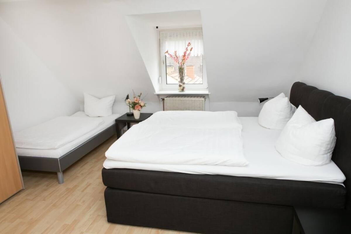 A bedroom with a large bed, a smaller bed, a nightstand with a vase of flowers, and a window with a view of the outside.