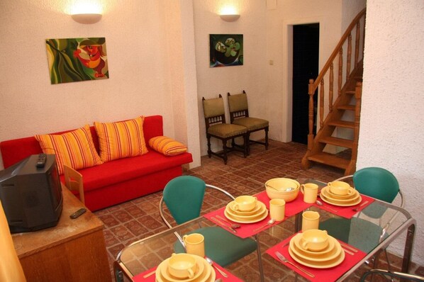 A living room with a red sofa, two armchairs, a glass dining table set with yellow dishes, and a wooden staircase.