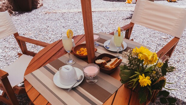 A round wooden table with a placemat, two glasses of a pink beverage, a white hat, a small bowl of fruit, a candle, and a vase with yellow flowers. - Antaira Bed and Tours (San Pedro de Atacama)