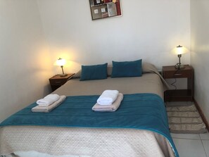 Una cama bien hecha con una colcha verde azulada y dos almohadas, dos lámparas de noche y una mesita de noche de madera con una lámpara.