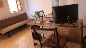A room with a wooden desk, a black monitor, a water dispenser, a brown sofa, and a window with sunlight. - Antaira Bed and Tours (San Pedro de Atacama)