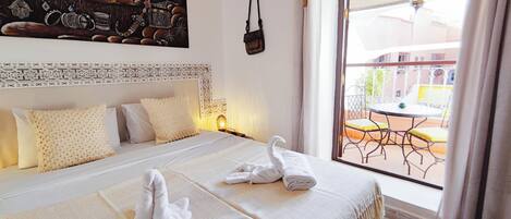 Habitación de hotel con cama, almohadas, mesita de noche, lámpara y vista al balcón exterior.