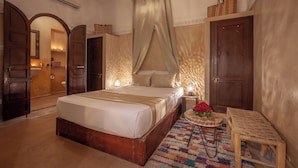 A bedroom with a large bed, a bedside table with a lamp, a small bench, and a door leading to another room. - Riad Kech Soul Boutique & SPA (Marrakech)