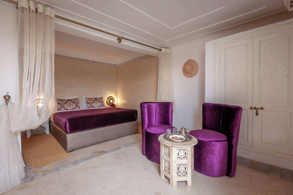Une chambre à coucher avec un lit, deux fauteuils violets, une petite table avec un plateau et une armoire.