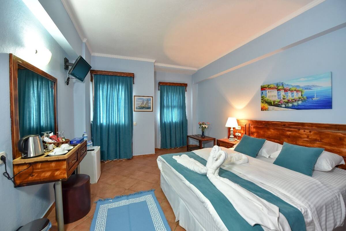 A hotel room with a bed, a desk with a kettle, a chair, a mirror, and a painting on the wall.