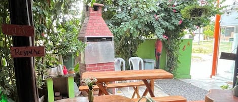 An outdoor patio with a round wooden table and green plastic chairs, a brick barbecue grill, and a green wall with a red chimney.