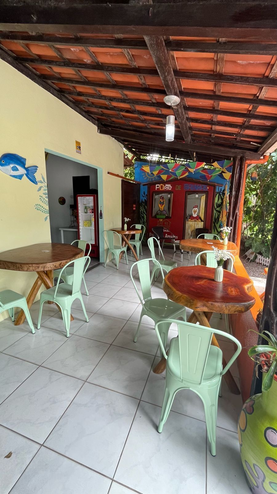 Un café al aire libre con mesas redondas y sillas verdes, piso de baldosas y un mural colorido en la pared.
