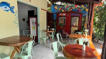 Un café al aire libre con mesas redondas y sillas verdes, piso de baldosas y un mural colorido en la pared.