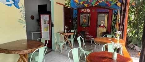 An outdoor cafe with round tables and green chairs, a tiled floor, and a colorful mural on the wall.