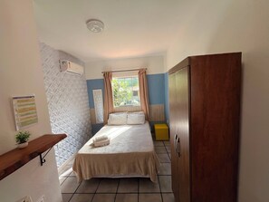 A bedroom with a bed, a bedside table with a plant, a wardrobe, and a window with curtains. - Pousada Porto de Amigos (Ipojuca)