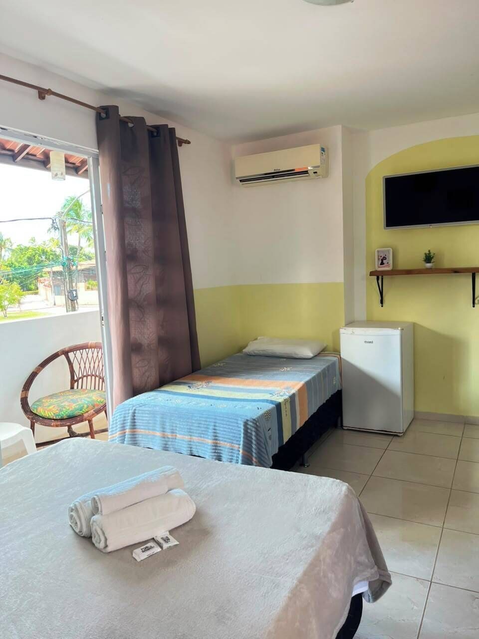 A room with a bed, a small refrigerator, an air conditioner, a chair, and a TV mounted on the wall.