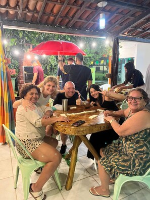 Un groupe de personnes est assis autour d’une table ronde en bois, un parasol rouge les protégeant du soleil, et elles sont en train de jouer à un jeu.