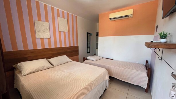 A hotel room with two beds, a mounted TV, an air conditioning unit, and a small shelf with a plant. - Pousada Porto de Amigos (Ipojuca)
