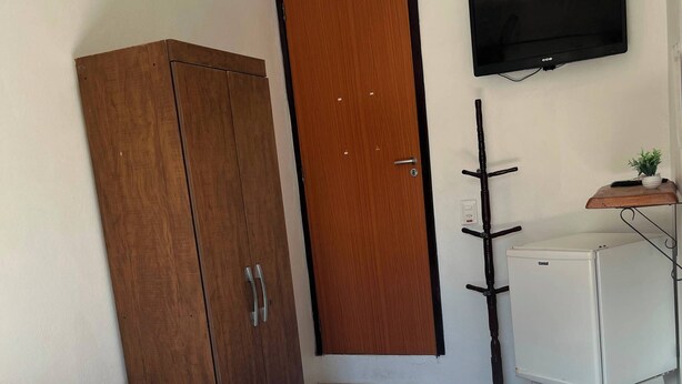 A bedroom with a bed, a wardrobe, a desk with a chair, a television, and a small plant. - Pousada Porto de Amigos (Ipojuca)