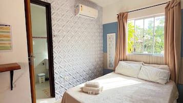 Habitación de hotel con cama, ventana con cortinas, puerta que da a un baño y un equipo de aire acondicionado en la pared.