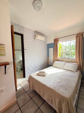 Una camera d'hotel con un letto, una finestra con tende, un ingresso per il bagno e un condizionatore d'aria a parete.