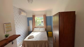 Quarto com cama, guarda-roupa de madeira, mesa de cabeceira com uma planta, radiador e janela com cortinas.