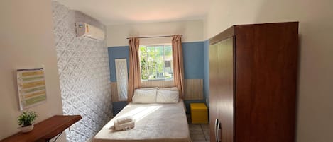Quarto com cama, guarda-roupa de madeira, mesa de cabeceira com uma planta, radiador e janela com cortinas.