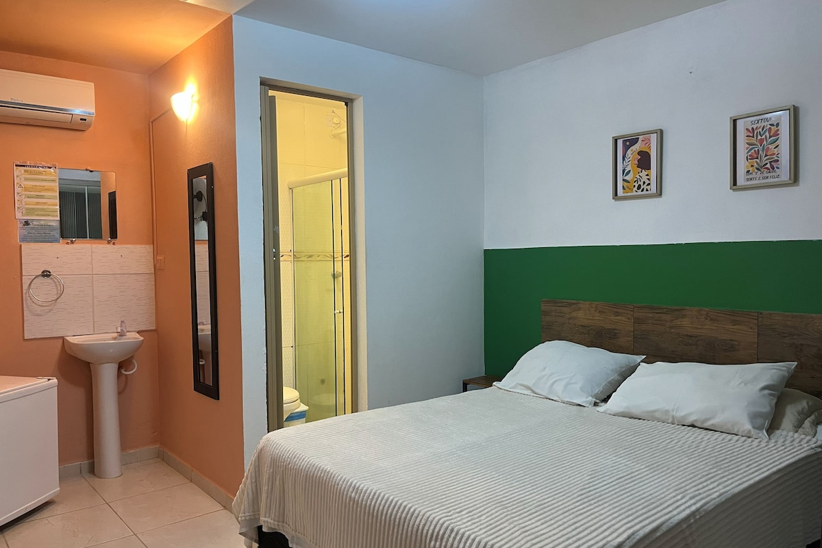A bedroom with a bed, a bedside table, a wall-mounted air conditioner, a mirror, and a door.