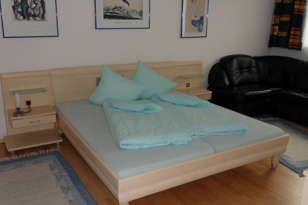 A neatly made bed with light blue bedding and pillows, a bedside table with a lamp, a black leather sofa, and framed artwork on the wall.